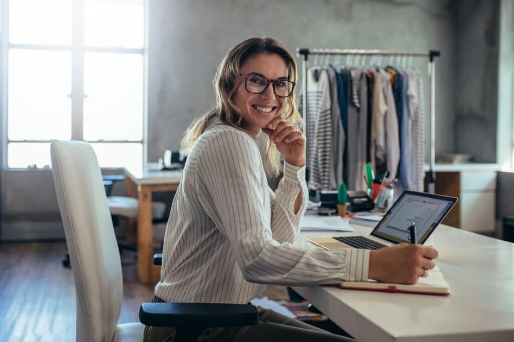 Empreendedora sorridente trabalhando com notebook em escritório de moda, fazendo anotações sobre emissão de nota fiscal eletrônica.